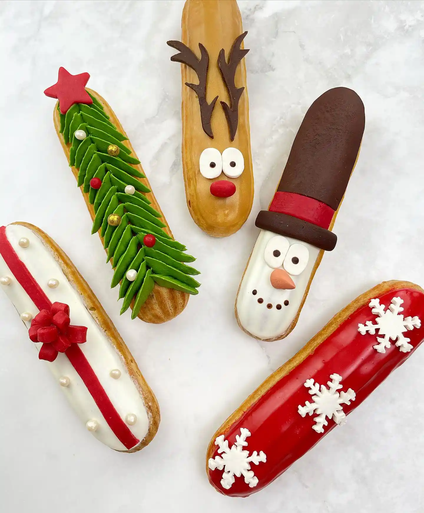 Five decorated Christmas eclairs on marble: a green tree with a red star, a reindeer, a snowman in a brown hat, a white one with a red bow and a red one with snowflakes